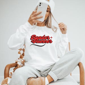 May include: White long-sleeve sweatshirt with the text "Reader + Runner" in red and black lettering. The text is in a retro font style.