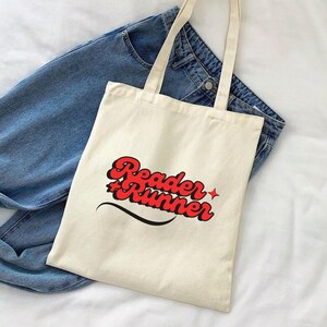 Puede incluir: Una bolsa de lona blanca con texto rojo y negro que dice "Reader Runner" en un estilo de fuente retro. La bolsa está sobre una superficie blanca con un par de pantalones vaqueros azules en el fondo.