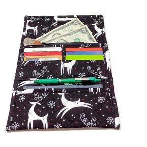 May include: Black fabric wallet with a festive reindeer and snowflake pattern. The wallet is open, revealing cash, credit cards, and a green pen. The wallet is quilted and has a white border.