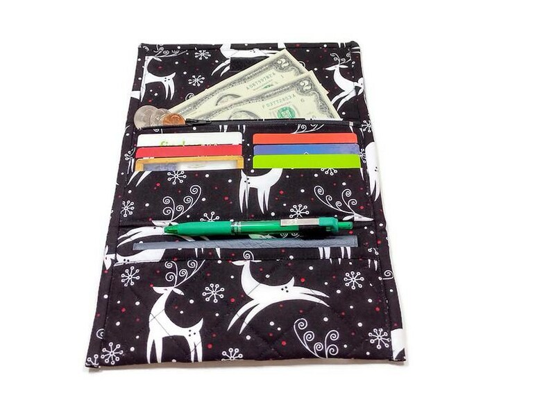 May include: Black fabric wallet with a festive reindeer and snowflake pattern. The wallet is open, revealing cash, credit cards, and a green pen. The wallet is quilted and has a white border.