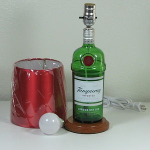 May include: A green glass bottle with a red label that reads "Tanqueray Imported London Dry Gin" is converted into a table lamp with a red lampshade and a white light bulb. The lamp is on a wooden base and has a white cord and plug.