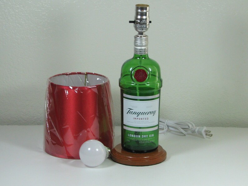 May include: A green glass bottle with a red label that reads "Tanqueray Imported London Dry Gin" is converted into a table lamp with a red lampshade and a white light bulb. The lamp is on a wooden base and has a white cord and plug.