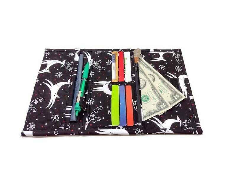May include: Black fabric wallet with a white deer and snowflake pattern. The wallet is open, revealing pens, credit cards, and a dollar bill. The wallet is designed to hold various items.