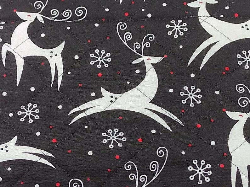 May include: Black quilted fabric with a repeating pattern of white reindeer and snowflakes, accented with small red dots. The reindeer have stylized antlers and are in various poses. A festive, winter-themed design.
