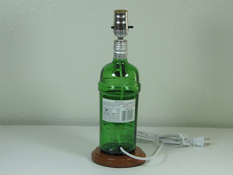 May include: A green glass bottle lamp with a wooden base and a white cord. The bottle has a label with the text "Bombay Sapphire" on it.