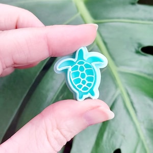 May include: A small, light blue and teal sea turtle charm featuring a raised, white-outlined shell pattern. The turtle has a smooth, matte finish and a distinct, symmetrical shape with four flippers and a head, designed for attachment to footwear.