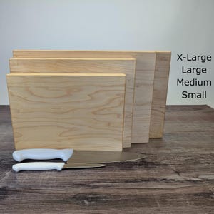 May include: A set of four wooden cutting boards in varying sizes: X-Large, Large, Medium, and Small. Two stainless steel knives with white handles are in the foreground. The boards are made of light-colored wood.