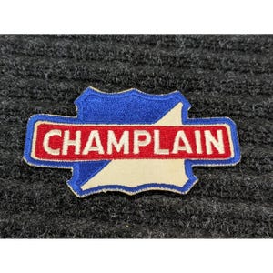 May include: A blue, red, and cream-coloured patch with the word "CHAMPLAIN" in red letters. The patch is in the shape of a shield with a blue border and a cream-coloured interior.