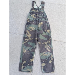 May include: Full-length denim overalls featuring a camouflage pattern in shades of green, brown, and black. The garment includes a front bib pocket with a small black rectangular brand label, adjustable shoulder straps with metal buckles, and side buttons.