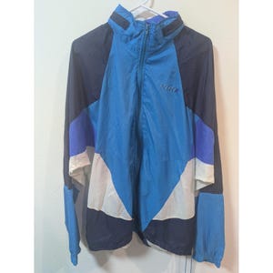 May include: A vintage Nike windbreaker jacket in shades of blue, navy, white, and purple. The jacket features a full zipper, a high collar, and the Nike logo on the chest. The color-blocked design adds a retro aesthetic to this athletic outerwear.