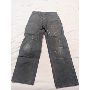 Könnte beinhalten: Dunkelgraue Arbeitshose mit mehreren Taschen und Kniebesätzen. Die Hose hat Farbspritzer in Weiß und anderen Farben, die Gebrauchsspuren aufweisen. Die Hose liegt flach auf einer weißen Oberfläche.