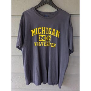 May include: A dark gray t-shirt with the word "MICHIGAN" in yellow block letters. Below the word "MICHIGAN" is a yellow block with the letter "M" and the numbers "1817". Below that is the word "WOLVERINES" in yellow block letters.