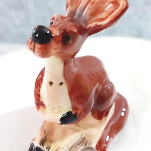 May include: A glossy ceramic figurine of a kangaroo in a seated position. The kangaroo is primarily reddish-brown with a cream-colored chest and belly, featuring a black nose, black paws, and black feet. It has large, upright ears.