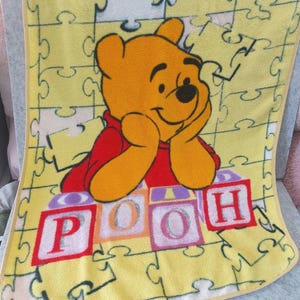 May include: Yellow baby blanket with a smiling Winnie the Pooh character in a red shirt. The blanket has a puzzle piece pattern and the word "POOH" in colourful blocks.