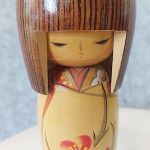 May include: A wooden Kokeshi doll with a brown, striped hairstyle and a painted face. The doll wears a yellow kimono with a floral design in red and white. The doll's head has two brown, rounded protrusions.