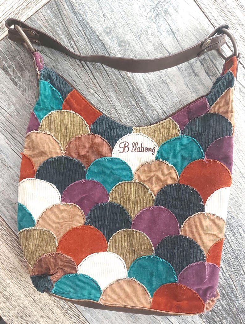 May include: A brown corduroy shoulder bag with a brown leather strap. The bag is decorated with a patchwork of colorful, scalloped shapes. The bag has a Billabong logo embroidered on the front.