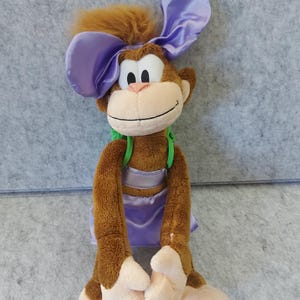 May include: A plush monkey toy with brown fur, large lavender ears, and a matching skirt. The monkey has a tan face, a pink nose, and a green harness. The toy is designed to sit upright.