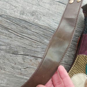 May include: A brown leather shoulder strap with two gold metal rivets. The strap is attached to a patchwork bag with various colors and textures.
