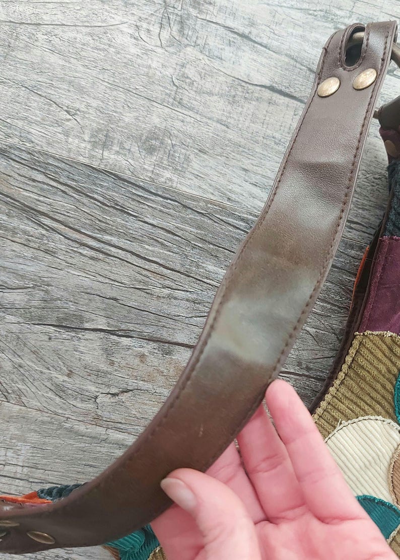 May include: A brown leather shoulder strap with two gold metal rivets. The strap is attached to a patchwork bag with various colors and textures.
