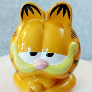 May include: A glossy ceramic figurine of the cartoon character Garfield, featuring a bright orange body with black stripes, white eyes with black pupils, a small pink nose, and a wide, light yellow muzzle, depicted in a seated position.