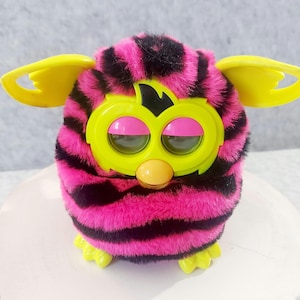 May include: A retro Furby toy with a pink and black striped body, yellow ears, and a yellow face with pink eyelids. The Furby has green eyes and a yellow beak. This electronic pet is a vintage collectable.