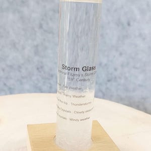 May include: A clear glass tube storm glass filled with liquid and white crystals, mounted on a square light-colored wooden base. The glass features black printed text including "Storm Glass," "Admiral Fitzroy's Storm Glass," and weather indicators.