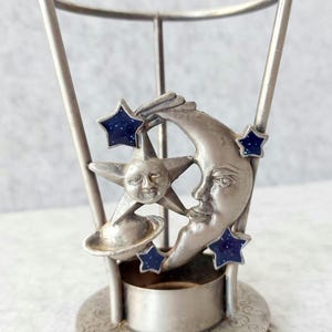 May include: A silver-toned metal candle holder featuring a celestial design with a crescent moon face, a star with a face, and a small planet. The structure includes three vertical supports and a circular base with embossed scrollwork patterns.