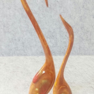 May include: Two abstract bird figurines crafted from a glossy, amber-toned material with swirling patterns of orange, yellow, and red. Each bird features a long, slender neck and a rounded base, standing upright with smooth, reflective surfaces.