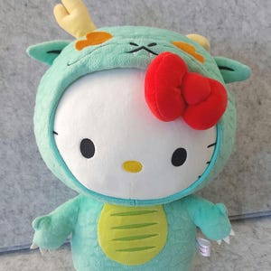 May include: A plush toy featuring a white character with black eyes and a red bow, dressed in a light green dragon costume with yellow accents. The costume includes a hood with horns and a belly plate.