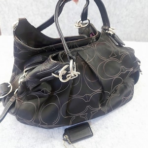 May include: Black handbag with a repeating pattern and silver hardware. The bag has two handles and a zipper closure. A small black tag and a charm with a carriage design hang from the bag.