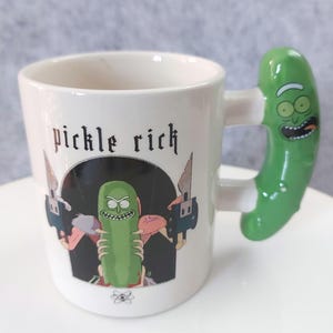 May include: White ceramic mug with a green pickle-shaped handle. The mug features the text "pickle rich" above a graphic of a green pickle character holding weapons. The handle is a glossy green, and the mug has a white interior.