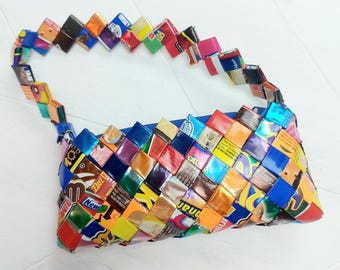 May include: A colorful woven handbag made from recycled candy wrappers. The bag has a blue strap and is decorated with a variety of colorful wrappers.