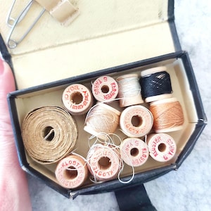 May include: A vintage sewing kit with a black case, open to reveal spools of thread in various colors, including white, black, and tan. The kit also contains a roll of tan ribbon, safety pins, and a small beige strap.