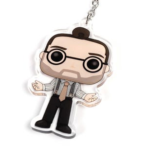 Könnte beinhalten: Ein Schlüsselanhänger mit einer Comicfigur mit einem schwarz-weiß gestreiften Hemd, braunen Haaren und einer Brille. Die Figur trägt eine braune Krawatte und Hosenträger.