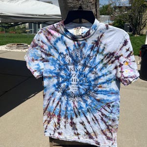 May include: A short-sleeved, tie-dye t-shirt in shades of blue, purple, and white. The shirt features a graphic with the text "KING GIZZARD ORCHESTRA" and "PHANTOM ISLAND TOUR". The shirt is hanging on a black hanger.