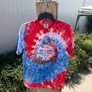 May include: A tie-dye t-shirt with a red, white, and blue spiral pattern. The shirt features the text "KING GIZZARD ORCHESTRA Phantom Island Tour" in a decorative emblem. The shirt is hanging on a black hanger.
