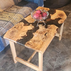 May include: A wooden coffee table with a black epoxy resin inlay. The table has a natural wood finish and a unique, organic shape. The table is sitting on a tiled floor and has a bowl of red apples on top.