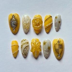 Puede incluir: Conjunto de diez uñas postizas amarillas y doradas con varios diseños, incluyendo un reloj, una mariposa, una rosa, un escudo y un pájaro.