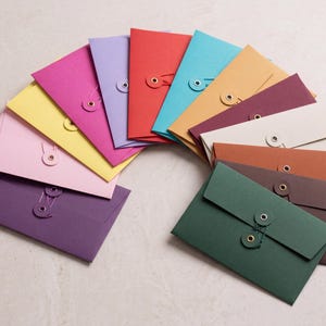 以下が含まれることがあります： 紫、ピンク、黄色、赤、青、オレンジ、クリーム、茶色、緑など、さまざまな色の長方形の紙封筒が扇状に並んでいます。各封筒には、紐とボタンの留め具が付いています。