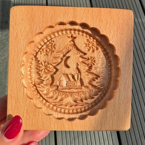 May include: A wooden cookie cutter featuring a detailed design of a Christmas tree, a church, and a crescent moon. The cutter is made from light brown wood and has a scalloped edge.