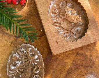 May include: A wooden cookie cutter with a bird and floral design, and a gingerbread cookie with the same design. The cookie cutter and cookie are on a wooden surface.