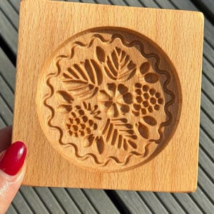 May include: A wooden cookie cutter with a detailed floral design. The cutter is round and has a scalloped edge. It is made of light brown wood.