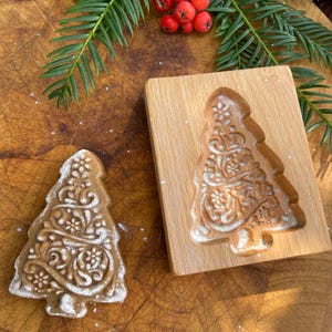 May include: A wooden Christmas tree cookie mould with a detailed design. The mould is on a wooden chopping board with a gingerbread cookie made from the mould.