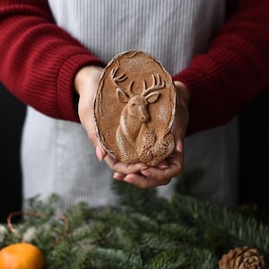May include: A brown, oval-shaped decorative plaque featuring a detailed relief of a deer with antlers and a pine cone. The plaque is held in front of a dark background, suggesting a rustic or holiday theme.