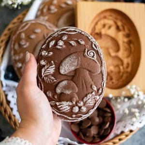 May include: A hand holds a brown gingerbread cookie with a mushroom design and white powdered sugar. The cookie is in front of a wooden cookie mold with a similar design.