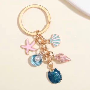 May include: A gold-colored keychain with multiple charms. The charms include a pink starfish, a blue seashell with a pearl, a pink and blue seashell, a blue seashell, and a white and blue seashell. The keychain is a beach-themed accessory.