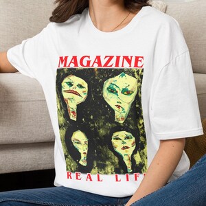 Peut inclure: Un t-shirt blanc à manches courtes avec une impression graphique représentant quatre visages stylisés dans des tons jaunes et verts sur un fond sombre. Le texte MAGAZINE apparaît en rouge au-dessus et REAL LIFE en rouge en dessous.