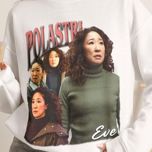Op de afbeelding: Een wit sweatshirt met ronde hals en een grafische print van het personage Eve Polastri. Het ontwerp bevat de tekst POLASTRI in een vetgedrukt rood lettertype bovenaan en de naam Eve in een wit scriptlettertype onderaan, met meerdere afbeeldingen.