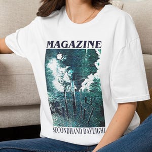 Peut inclure: Un t-shirt blanc à manches courtes avec une impression graphique portant le texte MAGAZINE en haut et SECONDHAND DAYLIGHT en bas. L'image centrale représente un paysage bleu sarcelle foncé avec un poteau et une forme de tête sur un ciel nuageux.