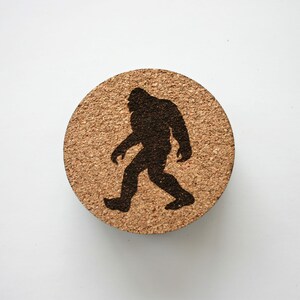 Puede incluir: Un posavasos redondo de corcho con una silueta negra de Bigfoot caminando hacia la derecha.